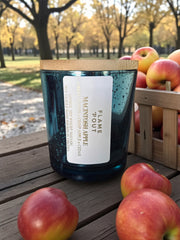 Candle with a wooden lid on a wooden table surrounded by apples in an outdoor setting